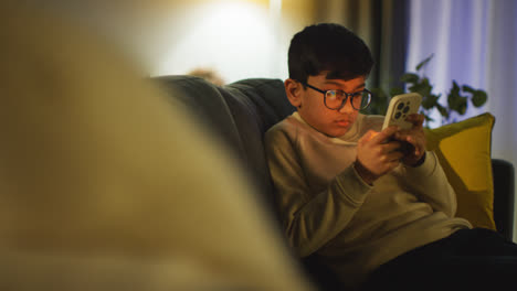 Niño-Sentado-En-El-Sofá-En-El-Salón-De-Casa-Jugando-En-El-Teléfono-Móvil-Por-La-Noche-3